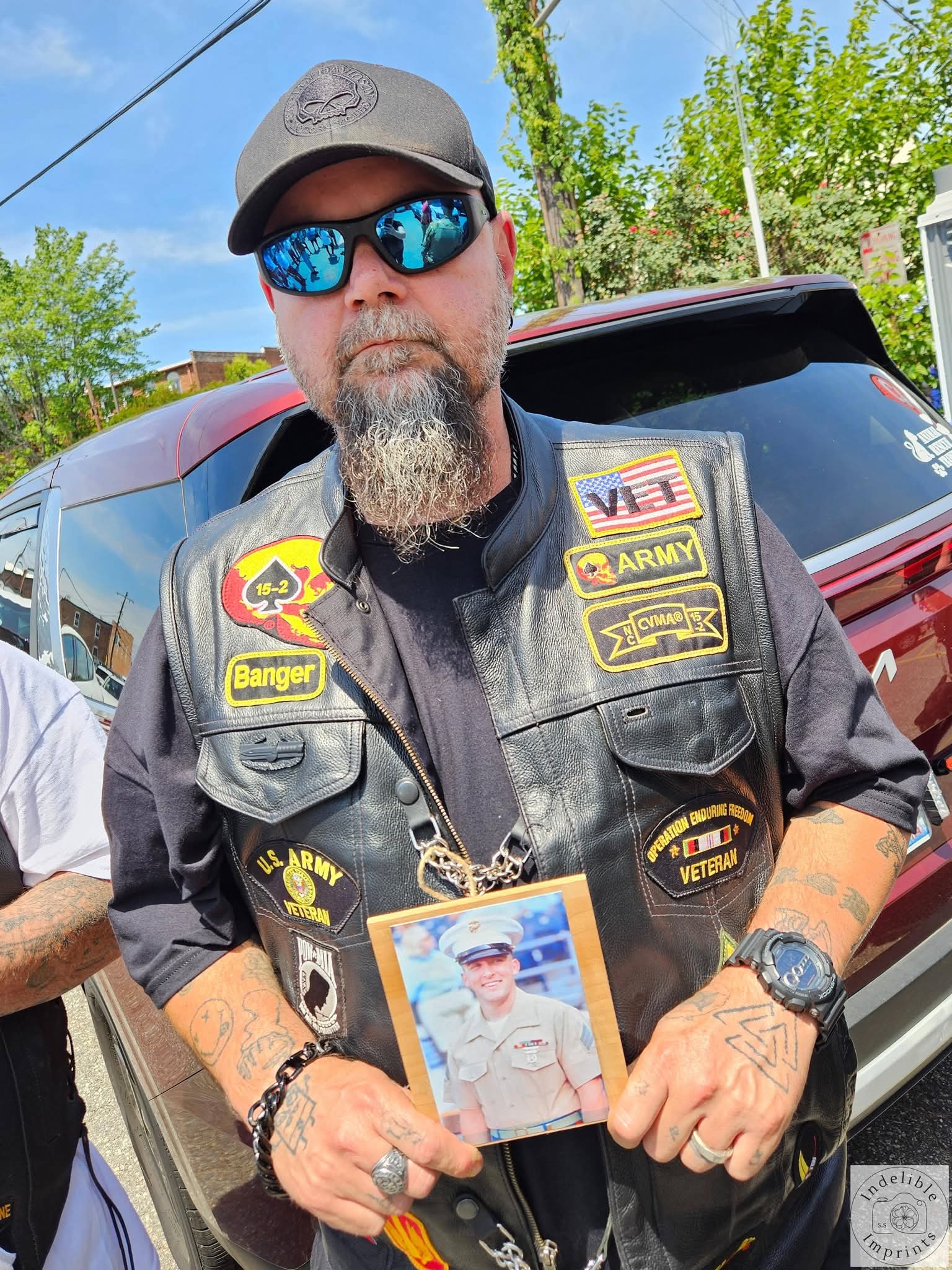 A veteran in a motorcycle club vest holding a memorial photo of a fallen Marine